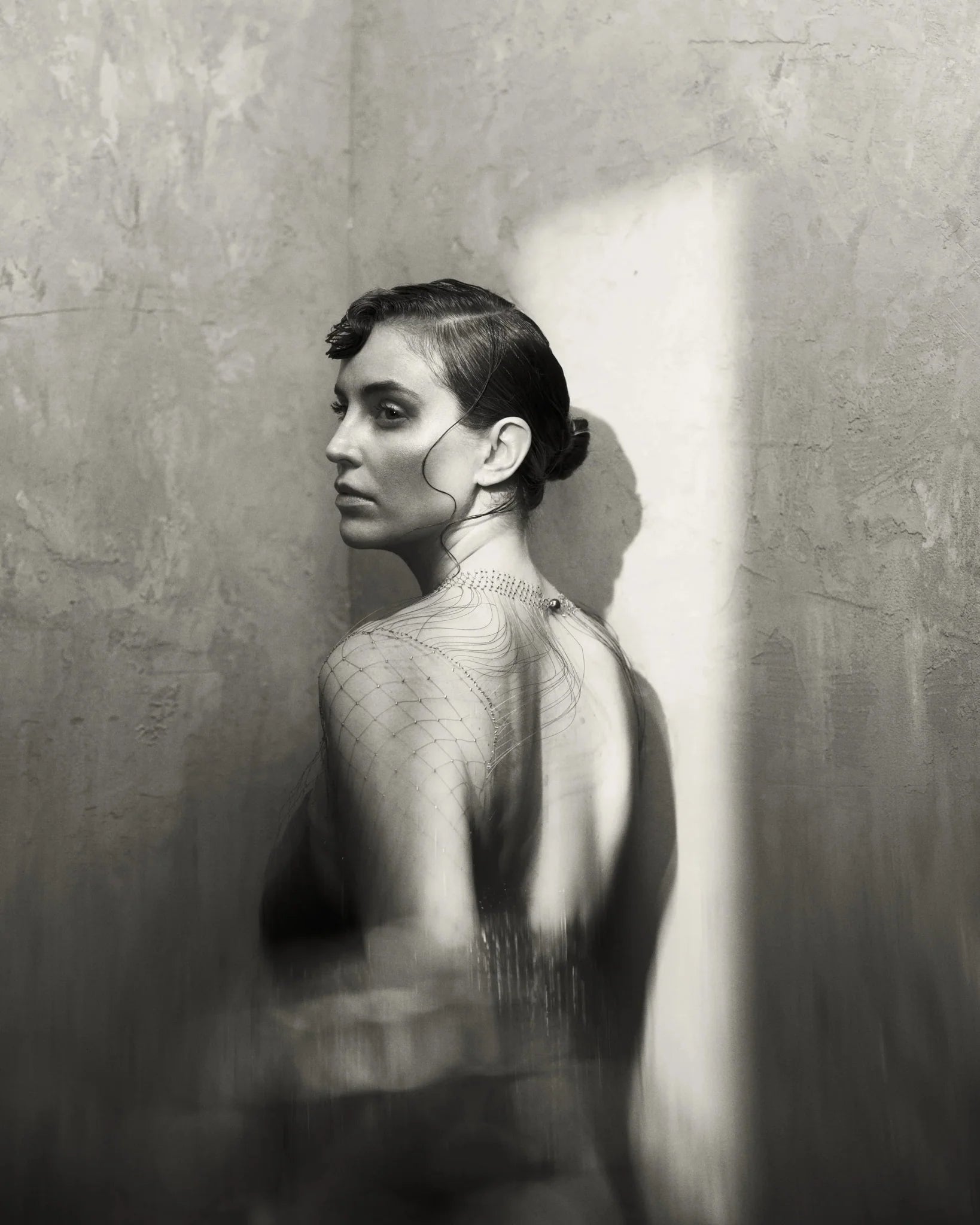 Black & white image with warm tones of a female model in a corner of a room with a window shaft of light projected from a Profoto Spot Small.