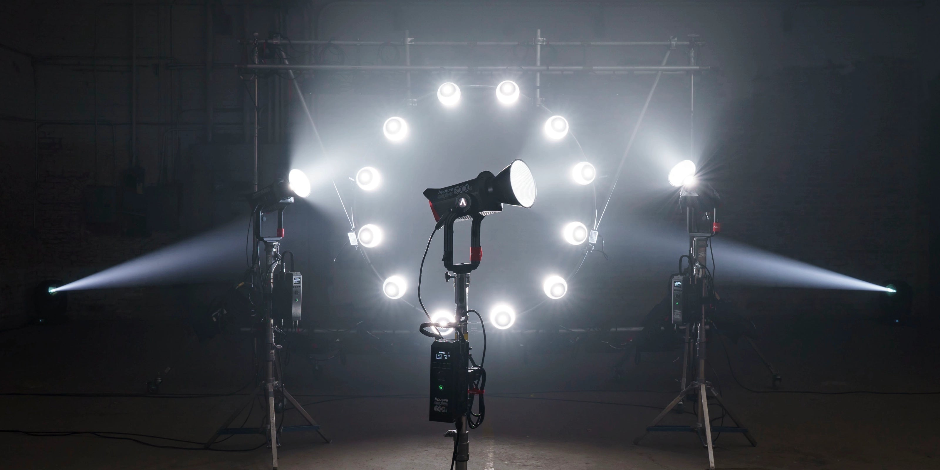 Aputure-LS 600d LED fixture and ballast on a stand, witha series of Aputure LS 600d lights forming a circle of light in the background, which is a warehouse type of scene. .