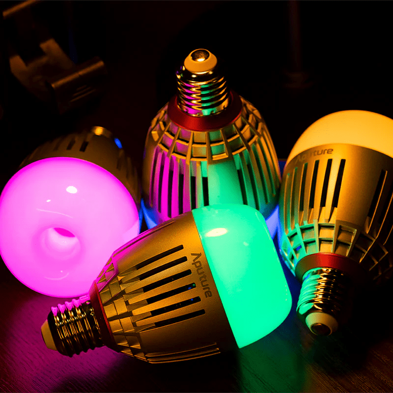 Four Aputure B7C full colour LED effect lamps, all at different colours of pink, green, yellow and blue sitting on a wooden table top and shown working via the intergrated battery.