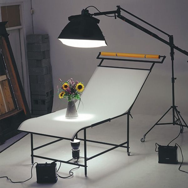 A Cambo ST-1 Frame for Shooting Table set up with lighting equipment, on wheels, against a studio background with a vase of flowers on top.