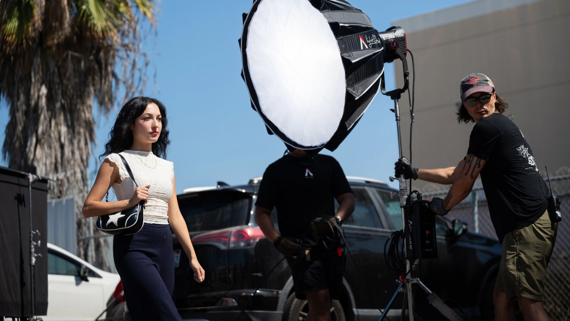 Film crew with a large Aputure 700x light setup outdoors, near vehicles and palm trees.