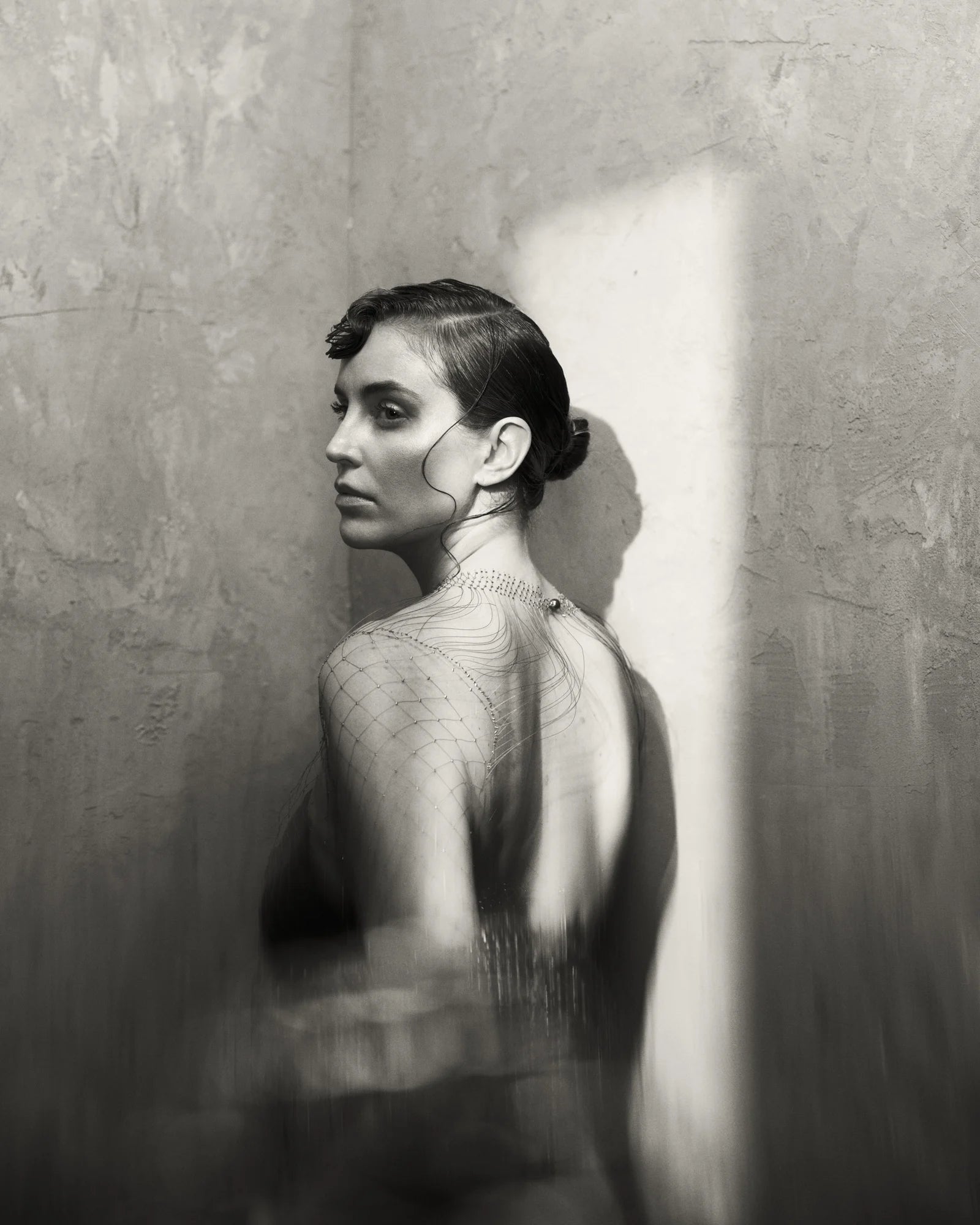 Black and white photo of a model against a rough wall, with shaft of wondow light projected onto her.